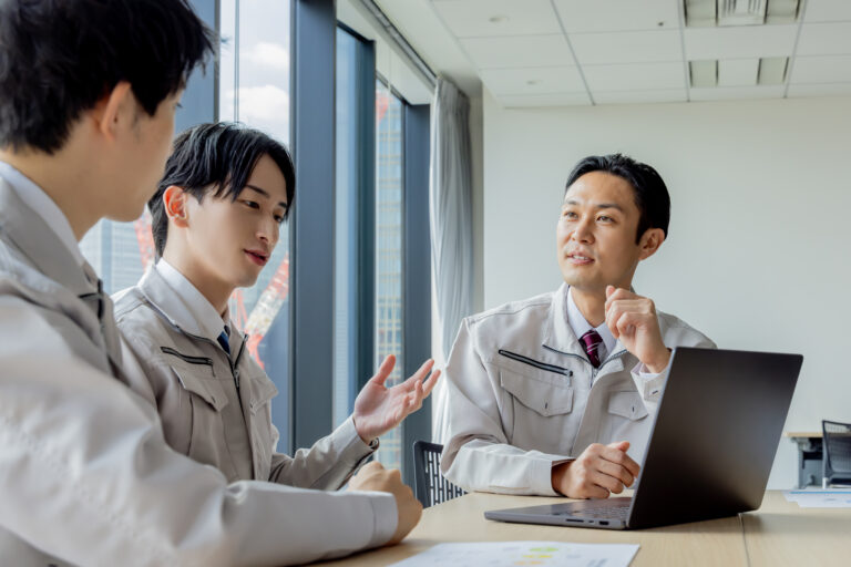 作業着姿の担当者3名がノートPCを囲み打合せする様子、窓の外にクレーンが見える